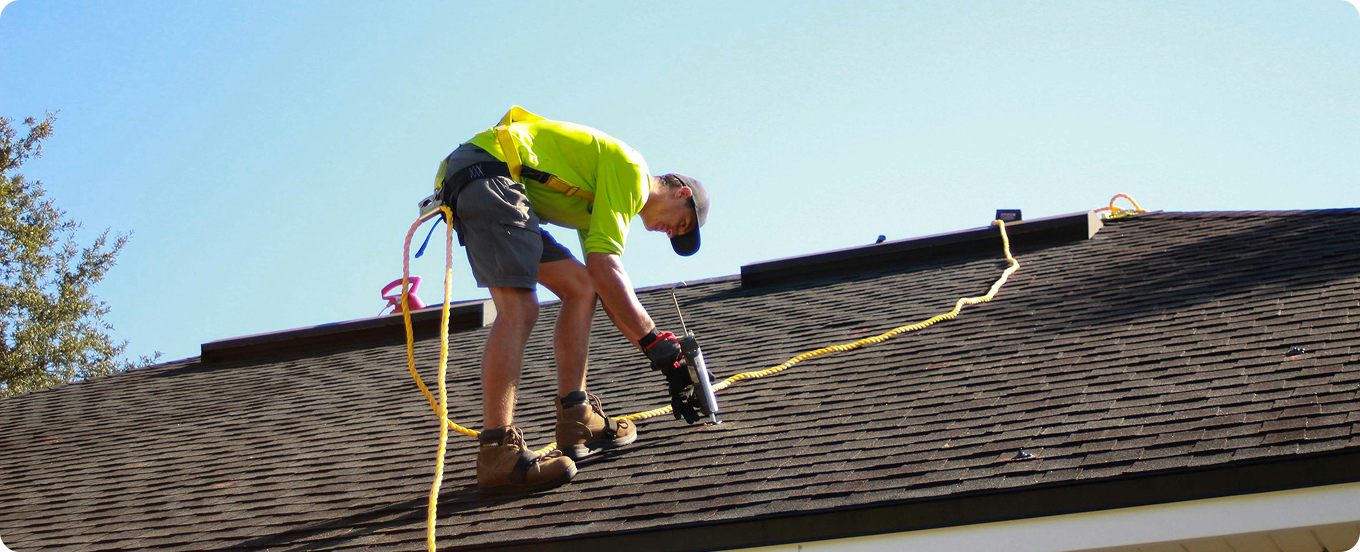 Roofing contractor performing maintenance and repairs using fall protection safety gear on a dark asphalt shingle roof.