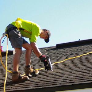 Roofing contractor performing maintenance and repairs using fall protection safety gear on a dark asphalt shingle roof.